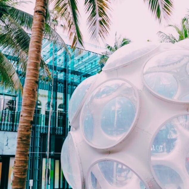 A white geodesic dome with large circular windows stands near palm trees and a modern glass building under a bright sky.