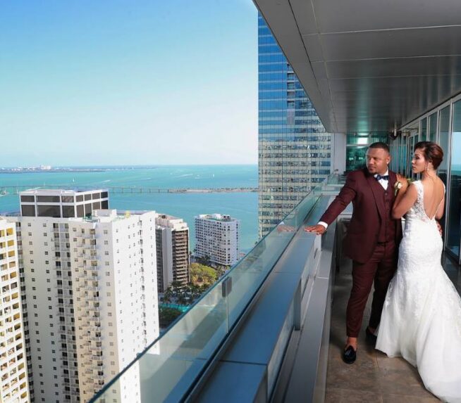 Bride and groom stand on a high-rise balcony overlooking a city skyline and ocean on a sunny day.