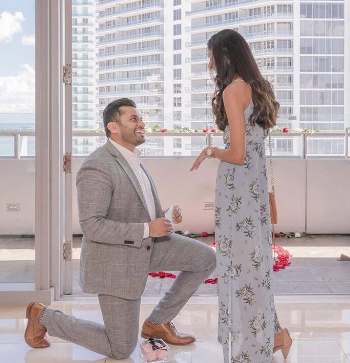 Man on one knee proposes to woman with ring box, surrounded by rose petals on a balcony.