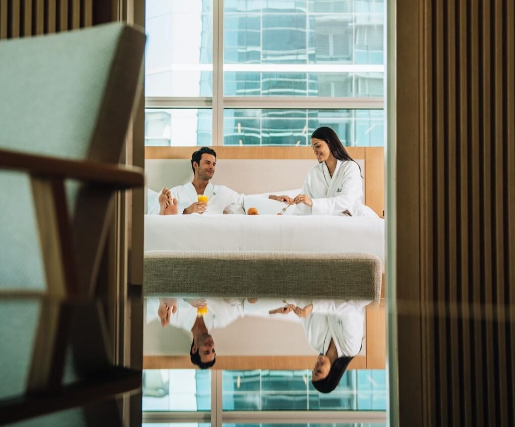 A couple in white robes sits on a bed, enjoying drinks, reflected on a glossy surface, with large windows and city buildings in the background.