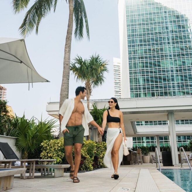 A couple in swimwear walks hand in hand by a pool, surrounded by palm trees and tall glass buildings on a sunny day.