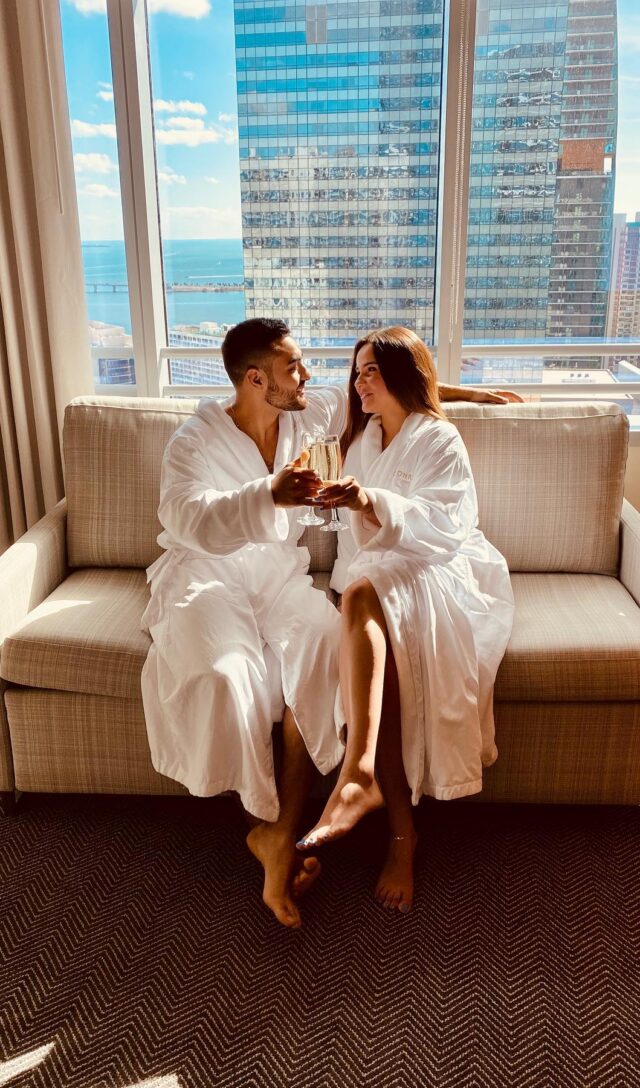 A couple in white robes sits on a sofa, toasting with champagne, with a city skyline view.