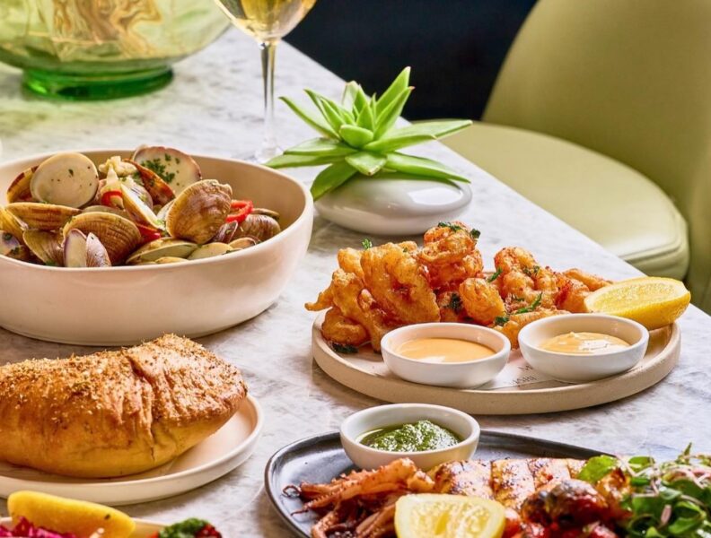 A table set with seafood dishes, grilled vegetables, bread, dipping sauces, a glass of white wine, and a small plant centerpiece.