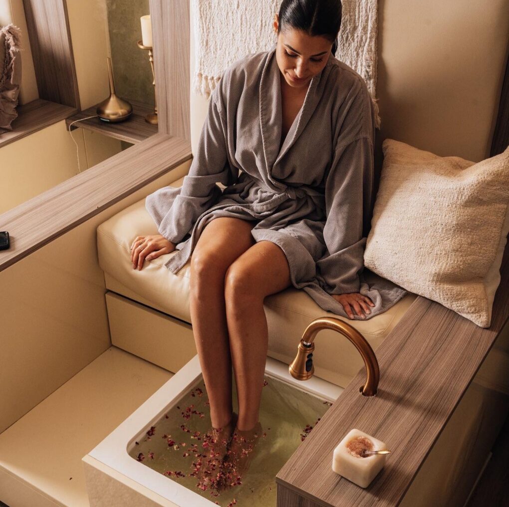 A woman in a gray robe enjoys a foot soak in a spa setting, with rose petals floating in the water and cozy decor around her.