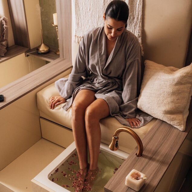 A woman in a gray robe enjoys a foot soak in a spa setting, with rose petals floating in the water and cozy decor around her.
