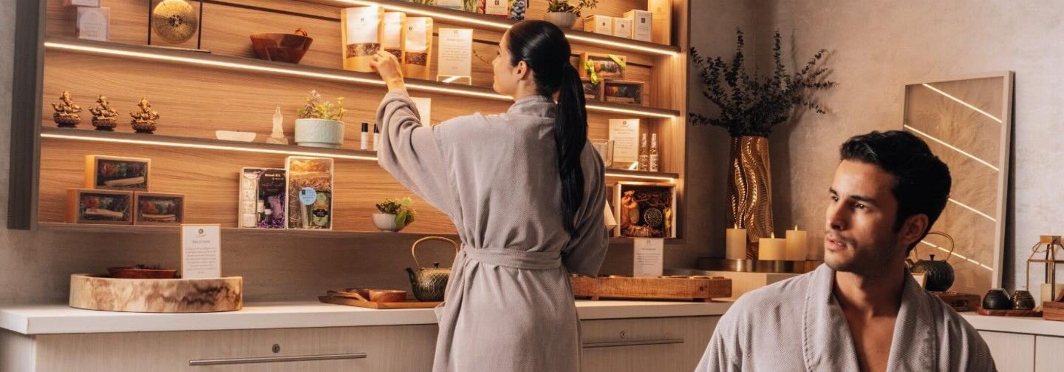 Two people in robes are in a modern, well-lit bathroom; one browses shelves of products, while the other sits on a bench with a green towel.
