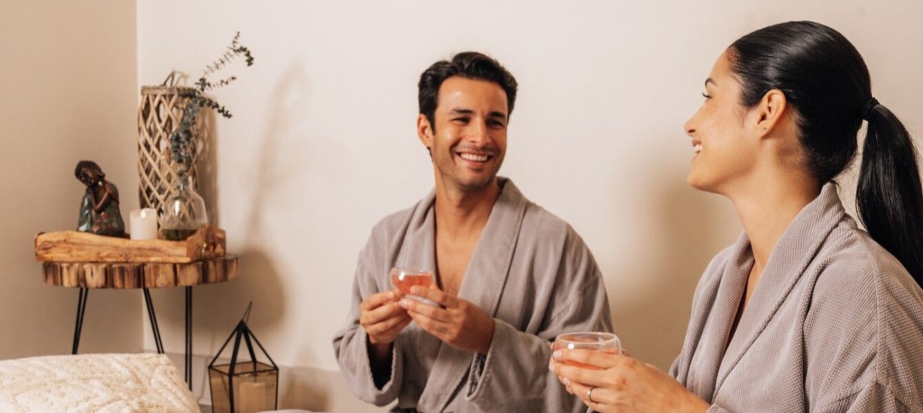 A man and a woman in gray robes sit on a cozy rug, smiling and drinking tea in a relaxing, softly-lit room with natural decor.