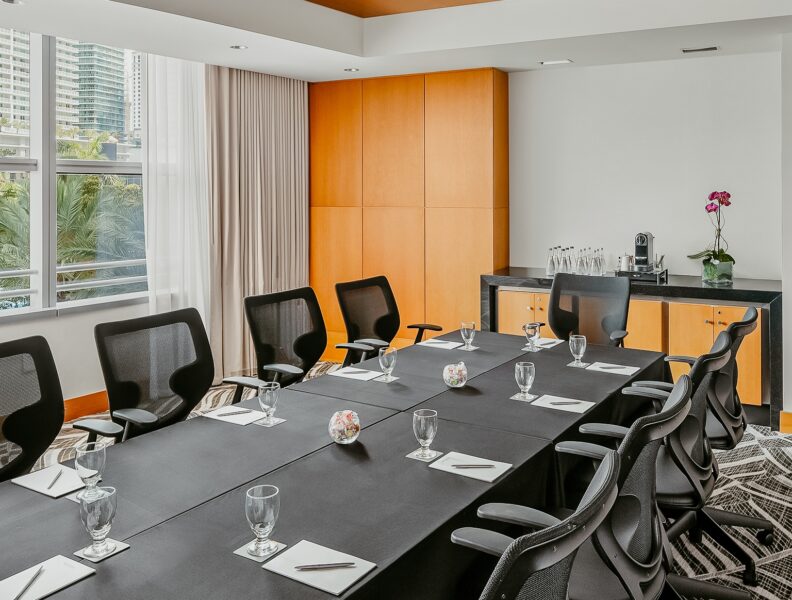 Modern hotel boardroom with a dark conference table, black mesh chairs, water glasses, notepads, warm wood ceiling panels, and city views through large windows.