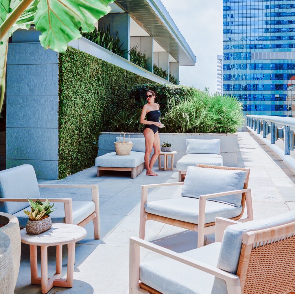 A woman in a black swimsuit stands on a sunny terrace with modern outdoor seating, lush plants, and tall city buildings in the background.