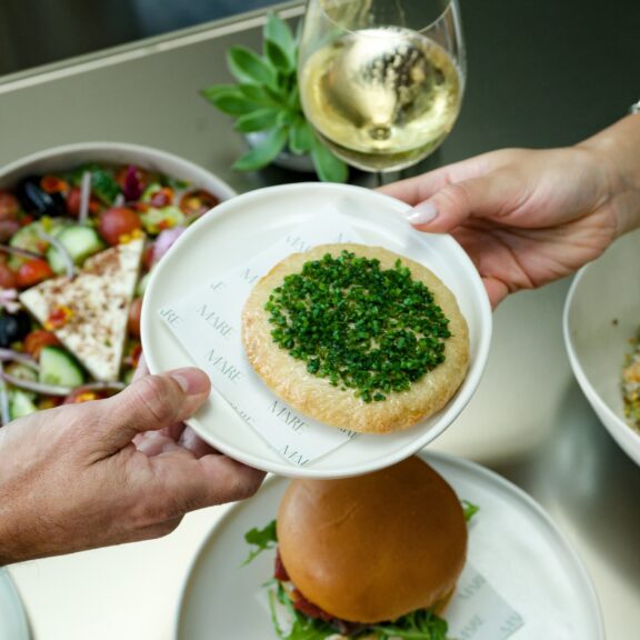 A person hands a plate of bread topped with chopped herbs to another, with wine, salad, and a sandwich visible on the table.