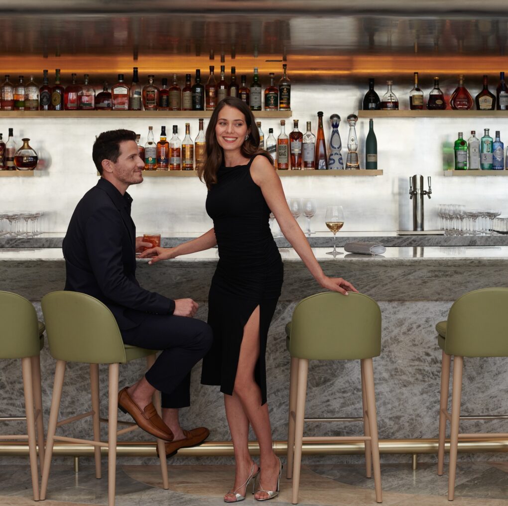 A man and woman in elegant attire sit and stand at a modern bar with bottles displayed behind them, smiling and enjoying drinks.