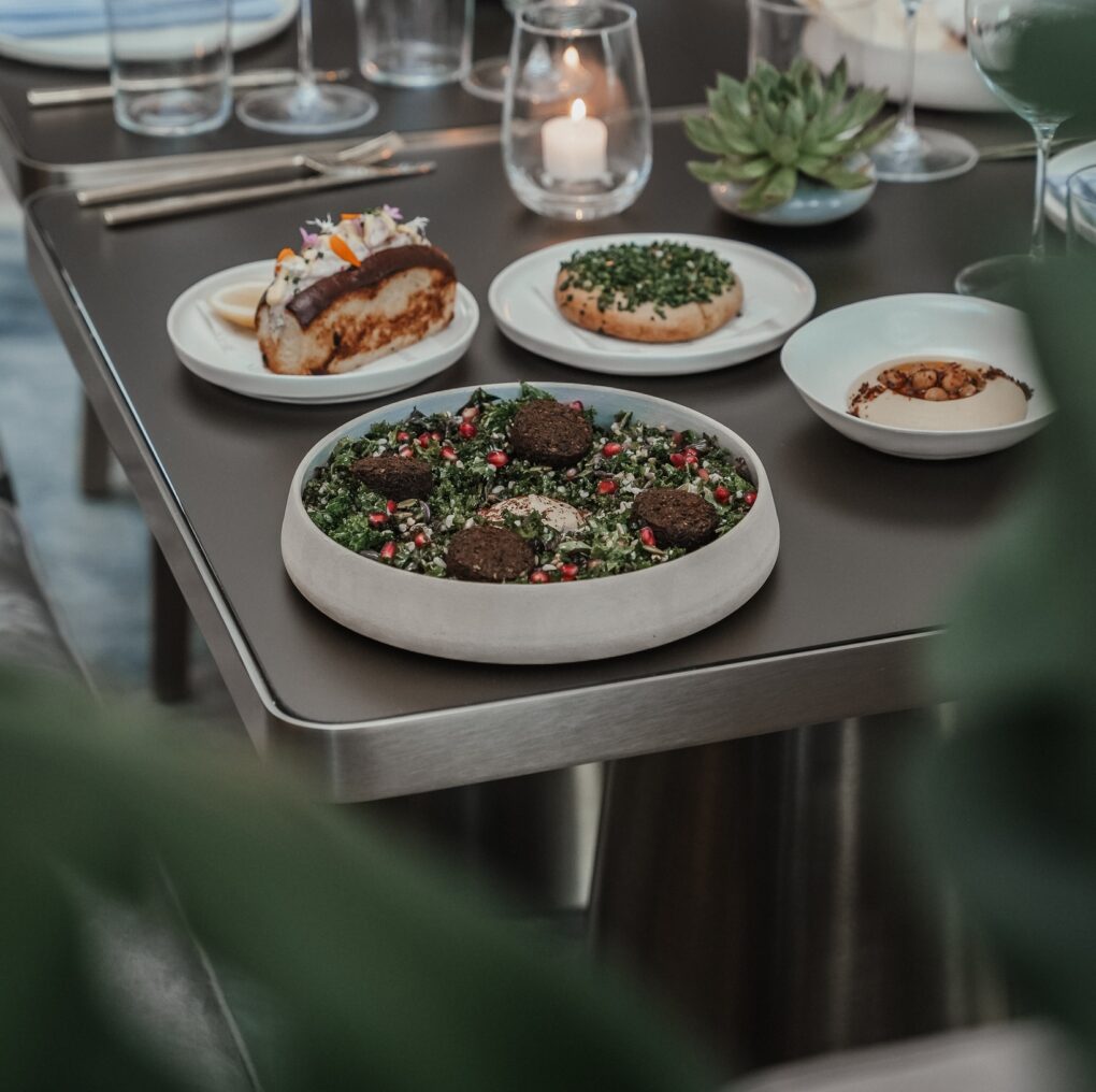 A table is set with a gourmet meal including a salad with falafel and pomegranate, a piece of fish with lemon, and a small bun with herbs.