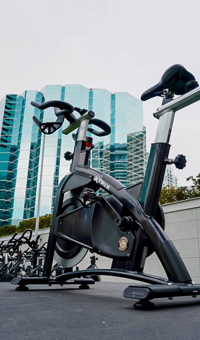 A stationary exercise bike is placed outdoors, with modern glass buildings and a cloudy sky in the background.