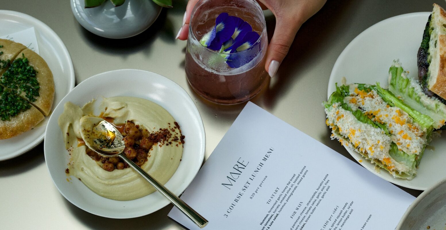 A hand holds a drink with purple flowers beside plates of partially eaten food, a menu, and a small succulent on a reflective table.