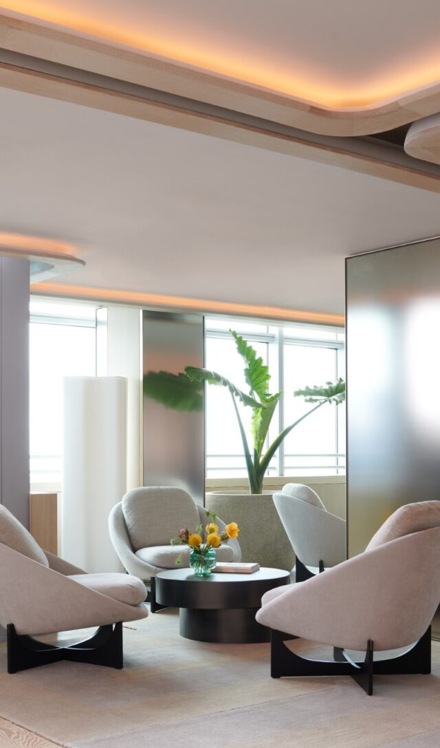 Modern lounge with four cream curved armchairs around a dark round coffee table, warm cove lighting, mirrored panels, and tropical plants by bright windows.
