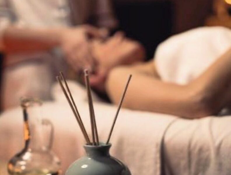 A vase with incense sticks and massage stones are in focus, while a person receives a massage on a table in the blurred background.