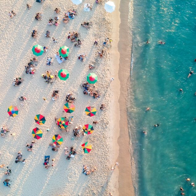 Aerial view of a crowded beach with colorful umbrellas on the sand and people swimming in the blue sea.