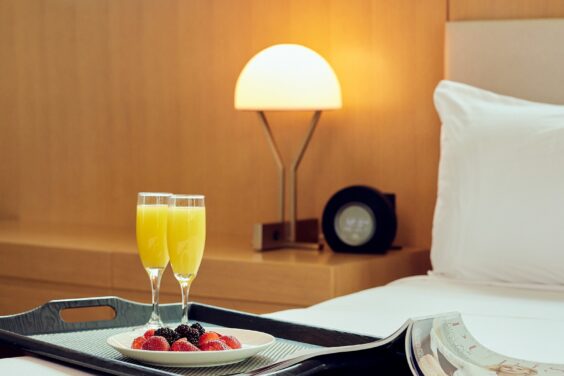 A tray with two glasses of orange juice, berries, and an open magazine on a neatly made bed in a modern hotel room.
