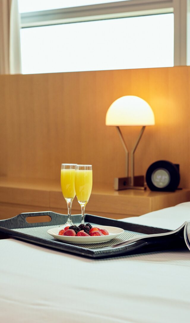 A tray with two glasses of orange juice, berries, and an open magazine on a neatly made bed in a modern hotel room.