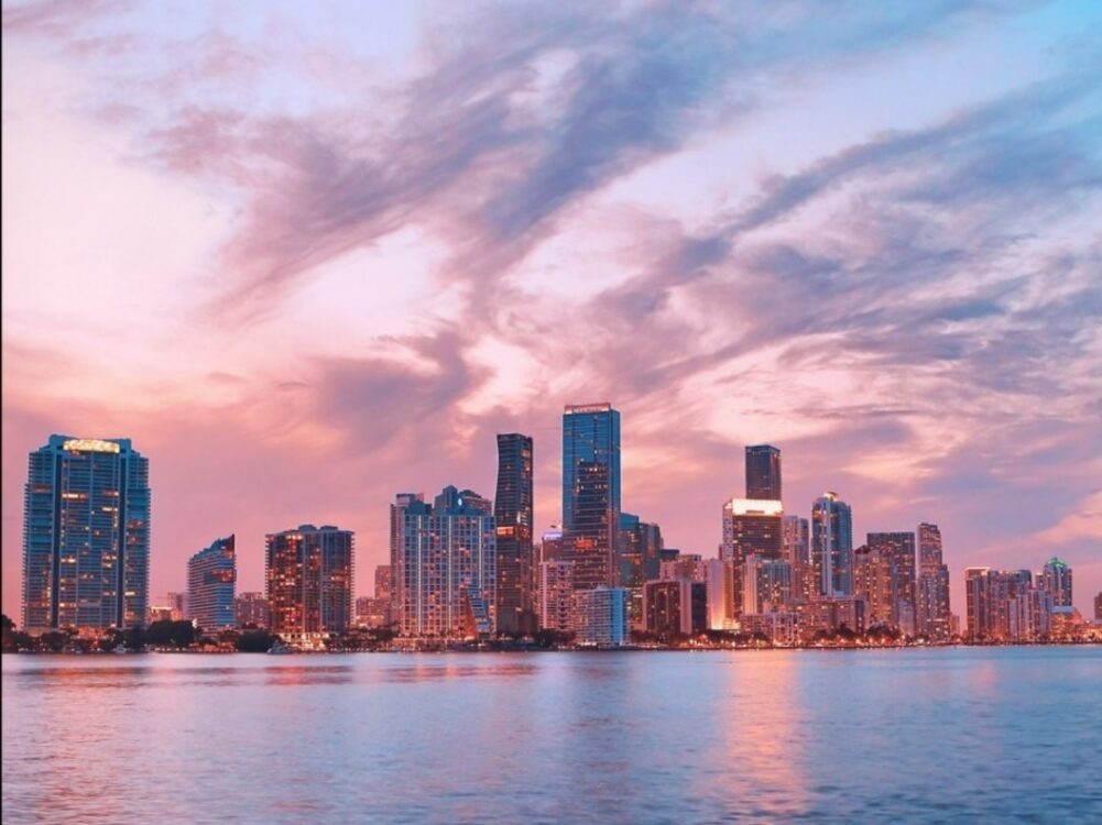 Miami skyline at sunset with tall buildings reflected in the water and colorful clouds filling the pink and blue sky.