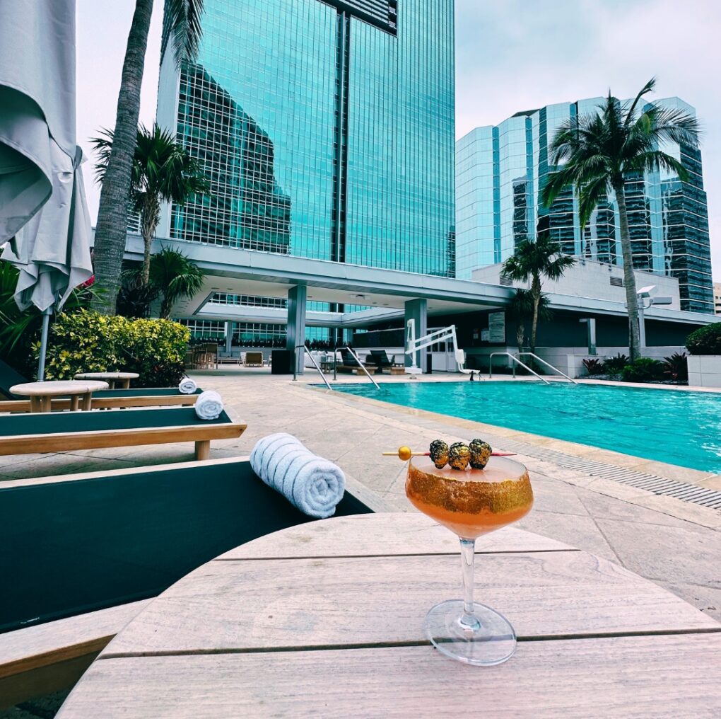 A cocktail sits on a poolside table with lounge chairs, palm trees, and tall glass buildings in the background.