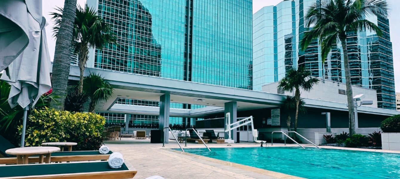 A cocktail sits on a poolside table with lounge chairs, palm trees, and tall glass buildings in the background.