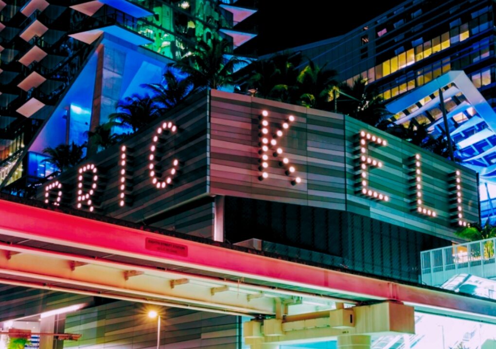 A brightly lit cityscape at night features modern buildings and a marquee sign with large illuminated letters. Palm trees and streetlights are visible.