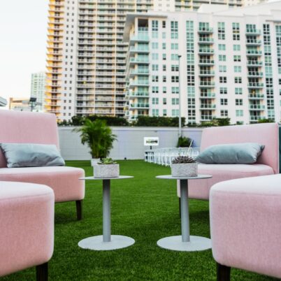 Outdoor seating area with pink chairs and small tables on green grass, set against a backdrop of modern high-rise buildings.