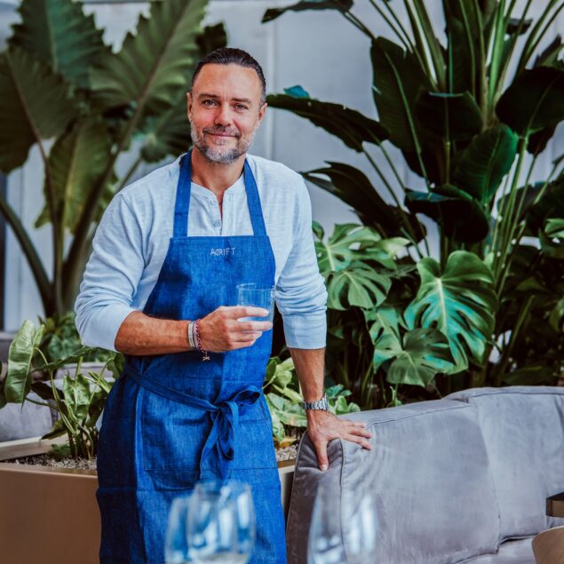 A man in a blue apron stands by a table holding a glass of water, surrounded by green plants in a modern, stylish indoor setting.