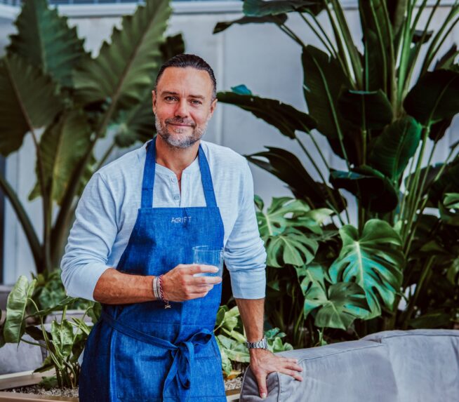 A man in a blue apron stands by a table holding a glass of water, surrounded by green plants in a modern, stylish indoor setting.