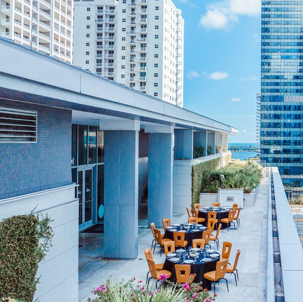 Outdoor rooftop patio with round dining tables and chairs, surrounded by tall modern buildings and greenery under a bright blue sky.