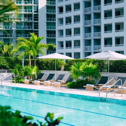 A modern outdoor pool with lounge chairs, white umbrellas, palm trees, and a tall apartment building in the background on a sunny day.