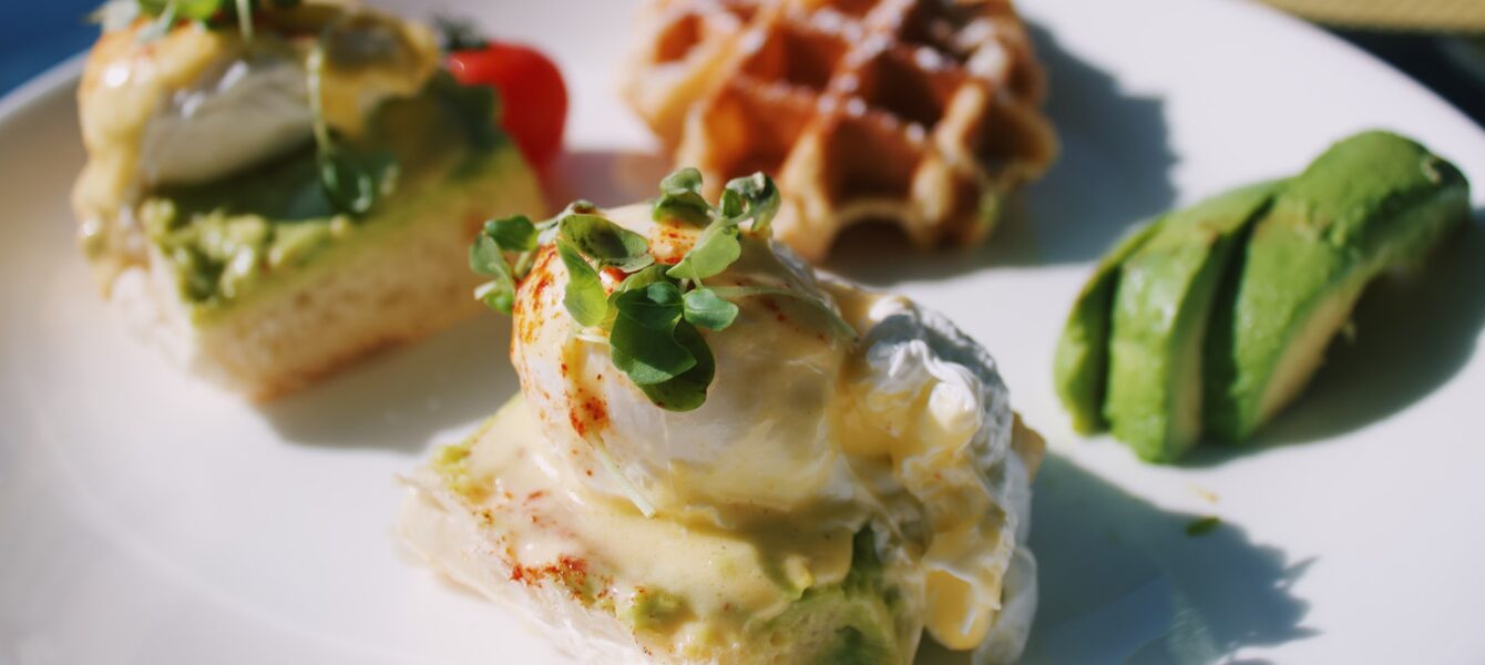 Two eggs Benedict with avocado and hollandaise sauce, a mini waffle, and sliced avocado on a white plate.