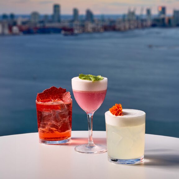 Three colorful cocktails on a white table with a city skyline and river in the background at sunset, under a sky with pink clouds.