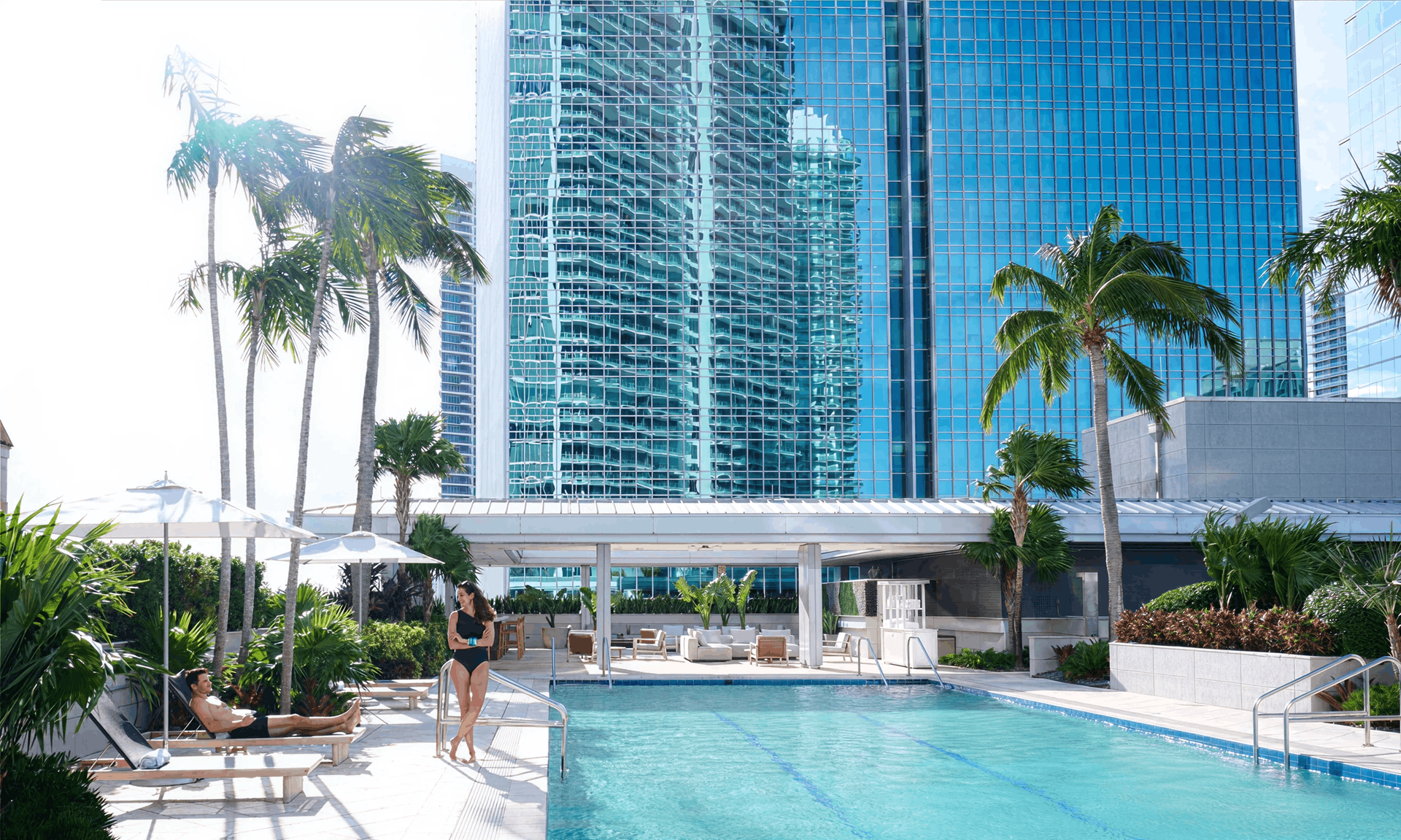 A man lounges and a woman stands by a pool, surrounded by palm trees and tall glass buildings under a clear sky.