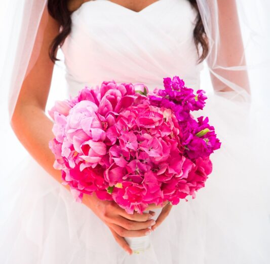 Bride holding a vibrant pink bouquet of peonies, hydrangeas and stock flowers, with a white veil and wedding dress.