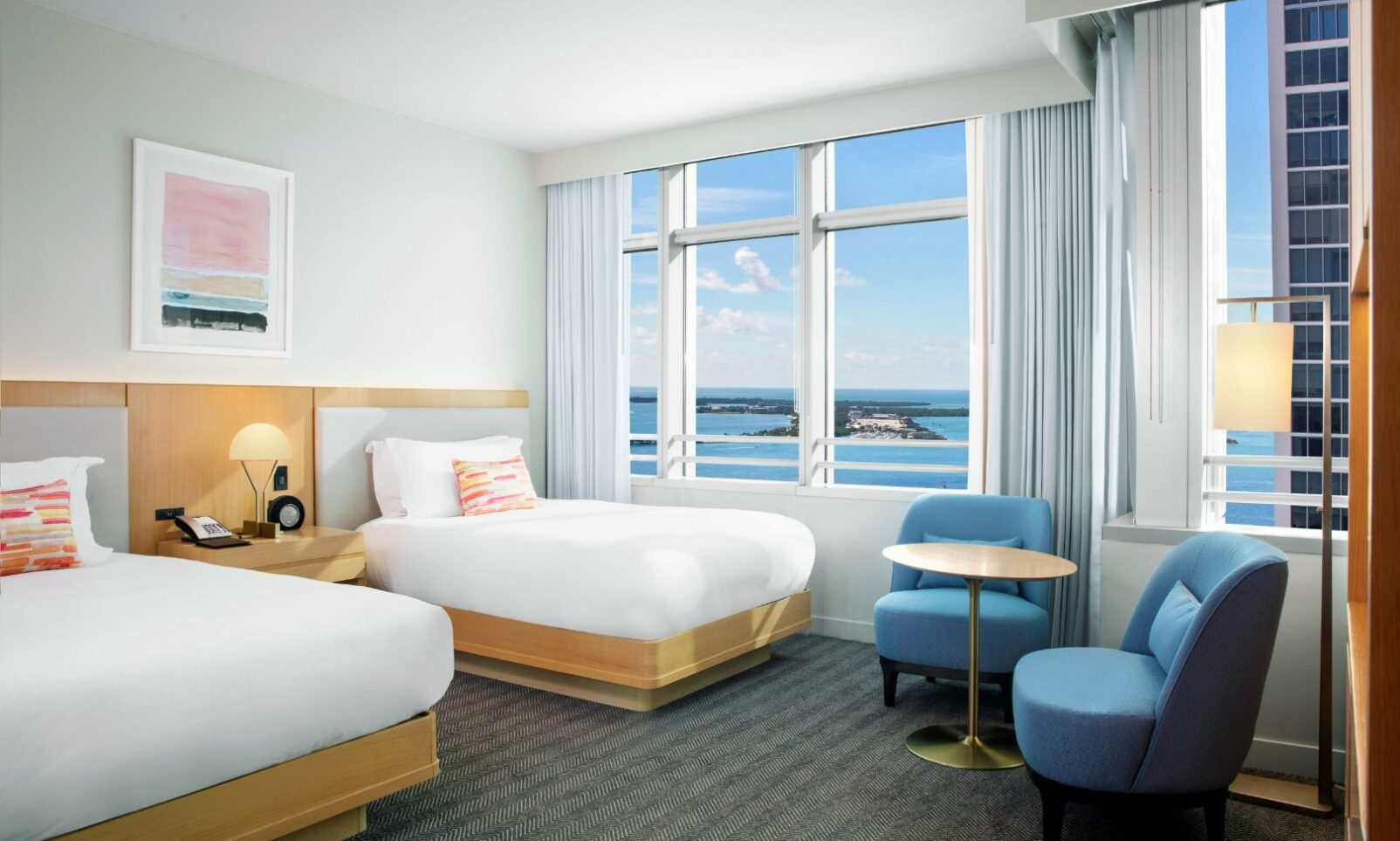 Modern hotel room with two beds, a small round table, two blue chairs, large windows, and an ocean view.
