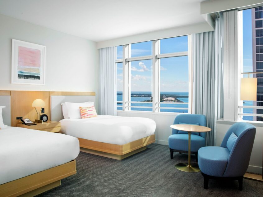 Modern hotel room with two beds, a small round table, two blue chairs, large windows, and an ocean view.