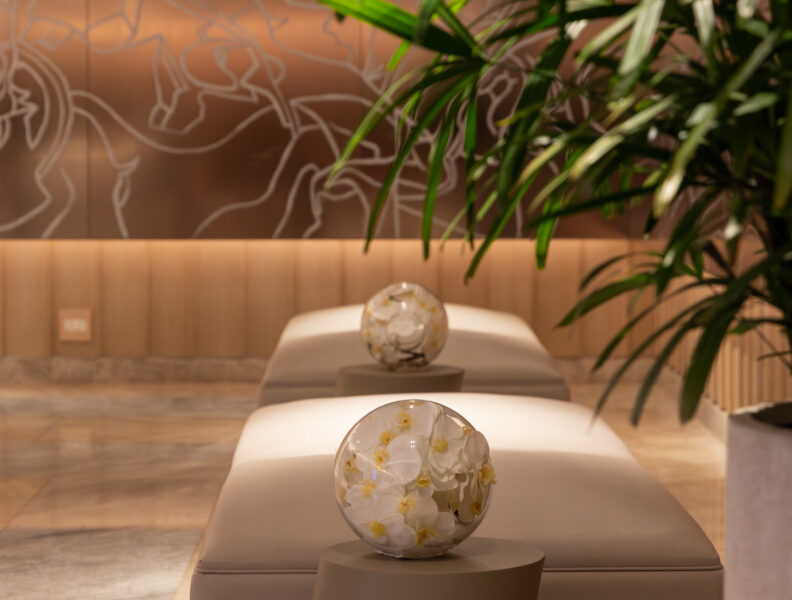 Modern lounge with beige benches, small round tables holding glass flower arrangements, and a large green plant in the foreground.