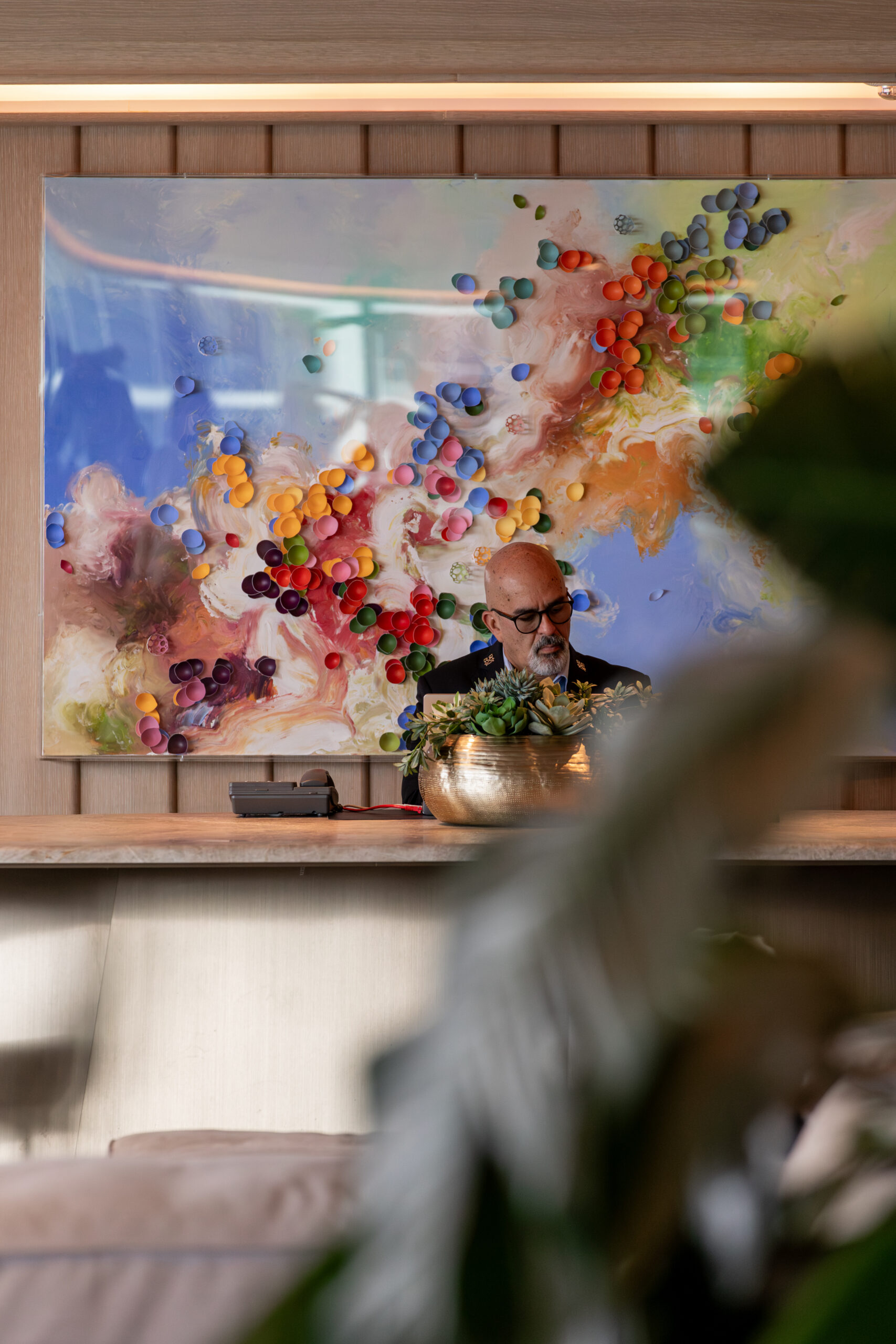 A man stands at a counter in front of a colorful abstract painting, with a plant in the foreground partially obscuring the view.
