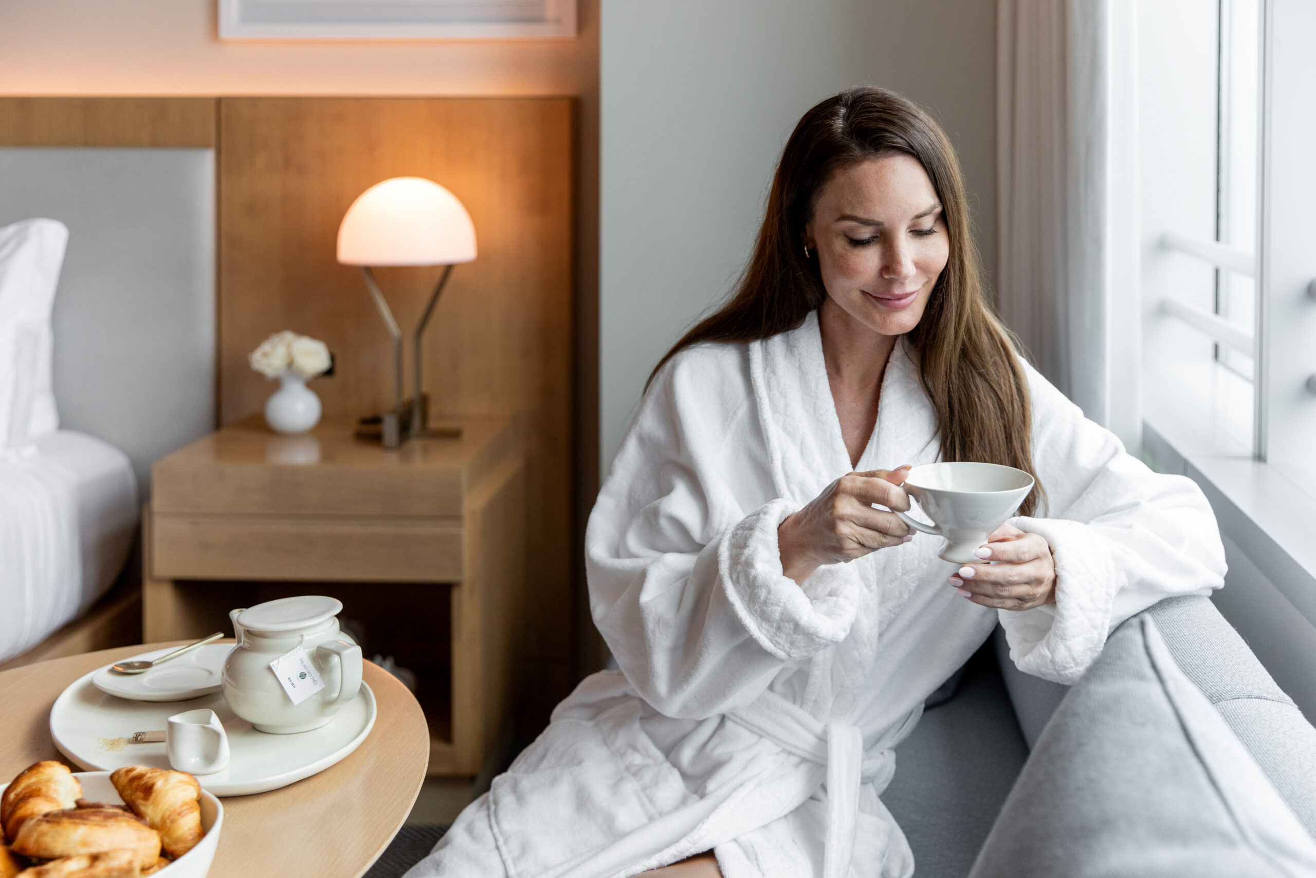Woman in a white bathrobe sits on a sofa by a window, holding a cup of tea, with breakfast and a teapot on a table nearby in a cozy room.