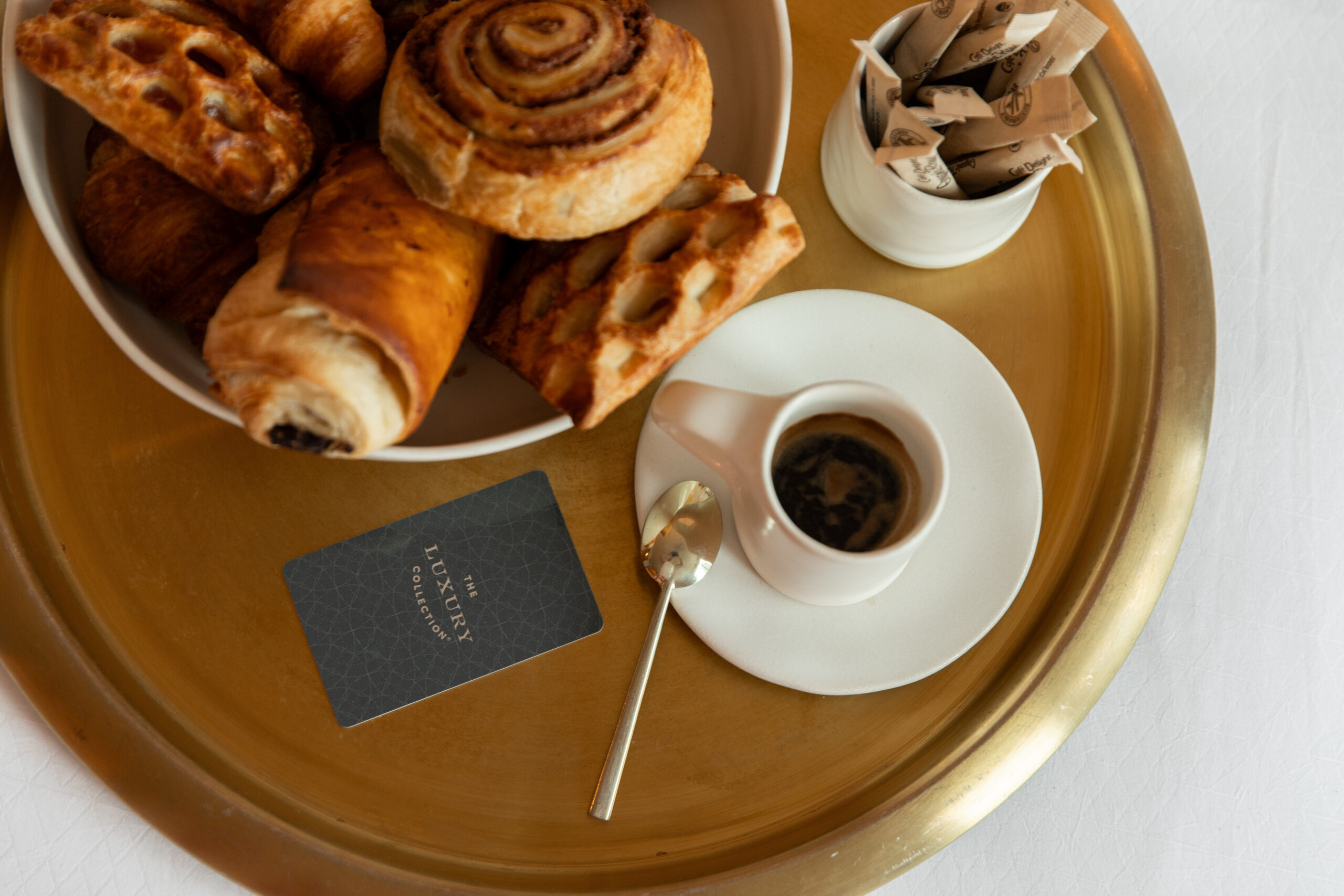 A gold tray with a cup of coffee, a spoon, a bowl of assorted pastries, sugar packets, and a card that reads The Luxury Collection.