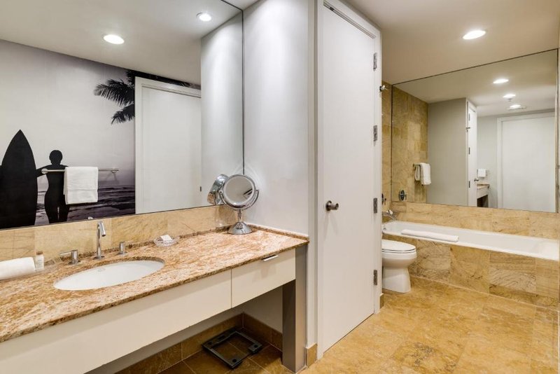 Modern bathroom with tan marble floor and countertop, large mirror, white sink, bathtub, toilet, and black-and-white surfer photo on the wall.