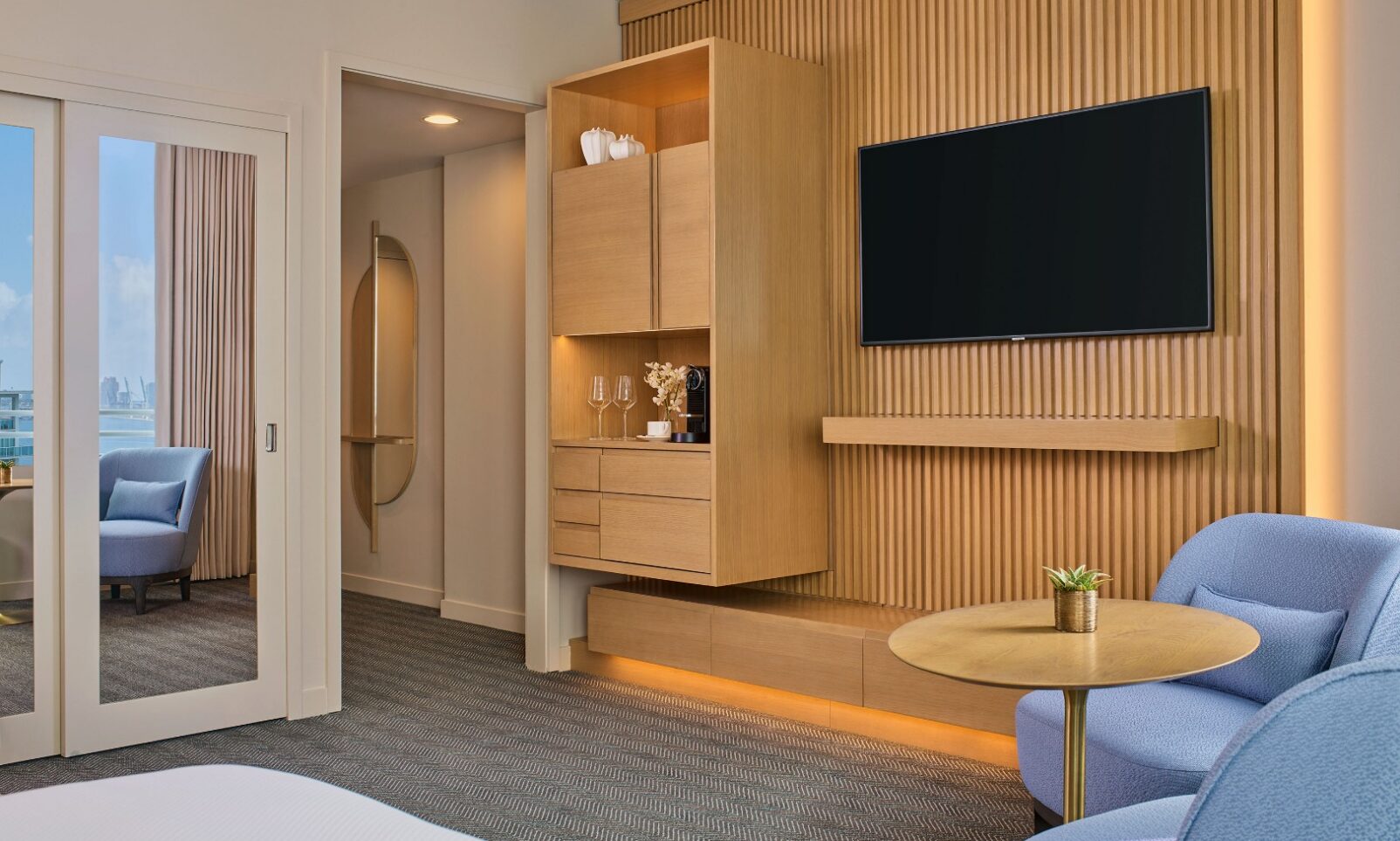 Modern hotel room with light wood furnishings, a wall-mounted TV, round table, blue chairs, and a sliding glass door opening to another room.
