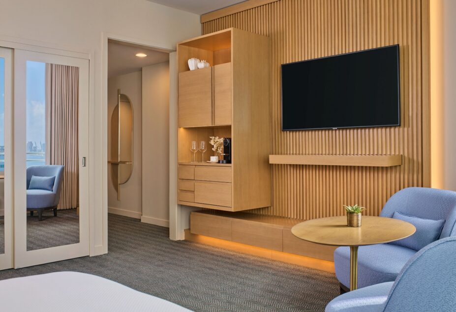 Modern hotel room with light wood furnishings, a wall-mounted TV, round table, blue chairs, and a sliding glass door opening to another room.