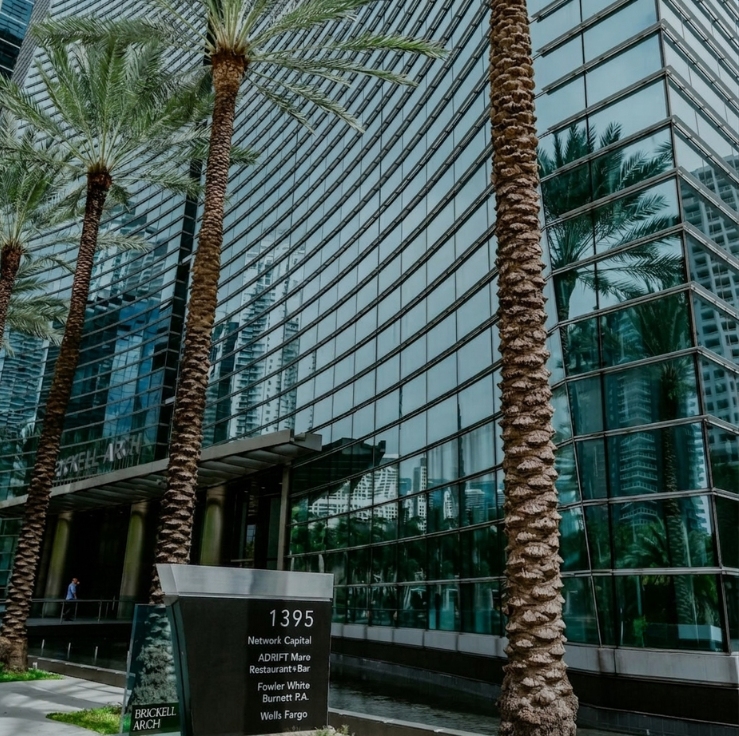 Modern glass office building with tall palm trees in front; a sign reads 1395 with business names listed below.