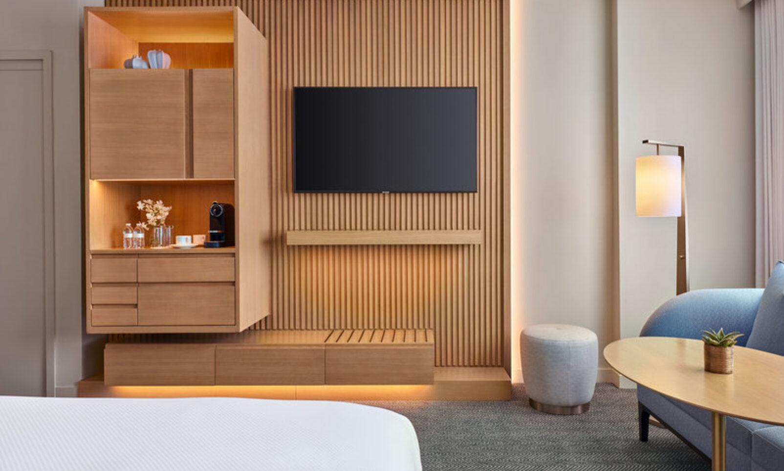 Modern hotel room with a wall-mounted TV, light wood paneling, open shelves with amenities, a blue sofa, round table, lamp, and large window.