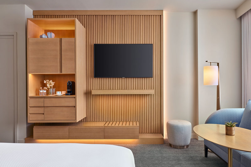 Modern hotel room with a wall-mounted TV, light wood paneling, open shelves with amenities, a blue sofa, round table, lamp, and large window.