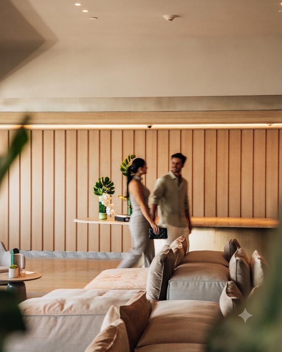 A couple walks hand in hand through a modern, stylish lounge with beige sofas and a wooden accent wall in the background.