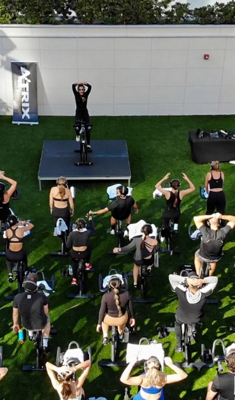 A group fitness class on stationary bikes outdoors follows an instructor on a stage, all raising their arms above their heads.